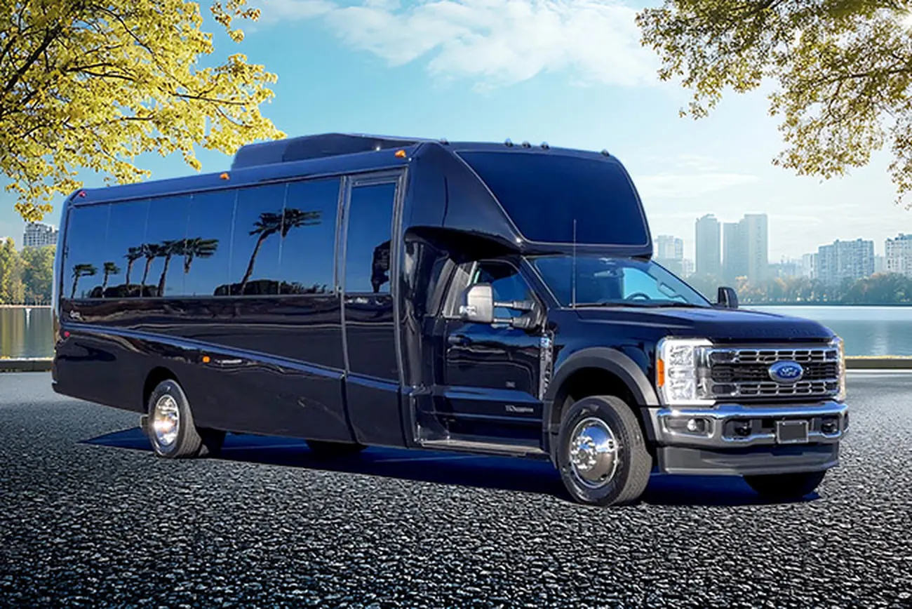 A dark 27-passenger Ford-based minibus parked beside a waterfront with trees and a city skyline behind it.