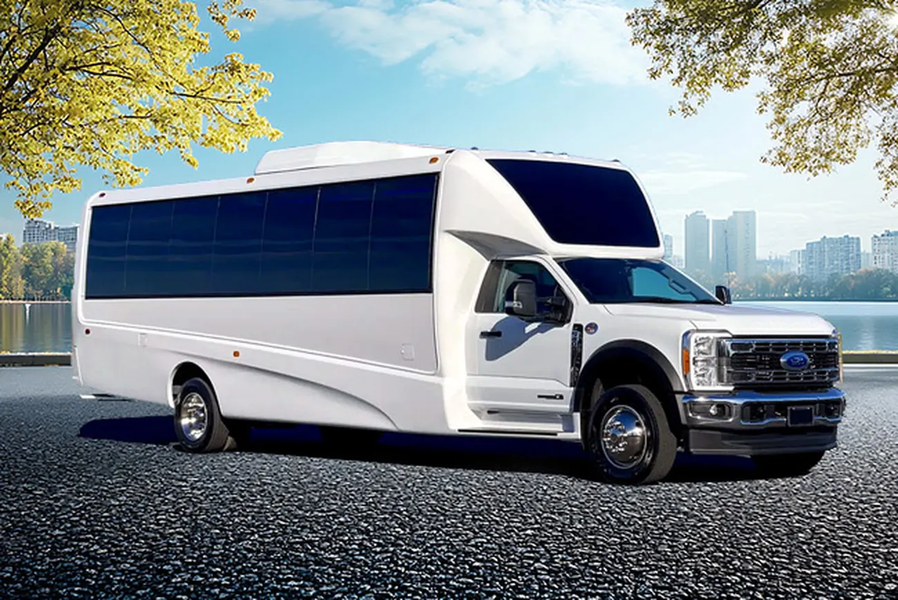A white 31-passenger Ford-based minibus parked beside a waterfront with trees and a city skyline behind it.